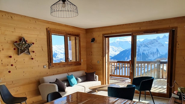 Un interior de madera con sofá, sillas, una mesa de comedor y ventanas amplias que ofrecen vistas a las montañas.