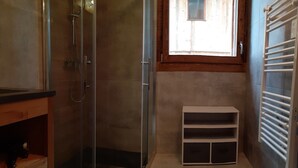 Un baño con cabina de ducha de vidrio, tocador de madera, estante blanco y ventana con vistas a una construcción de madera en el exterior.