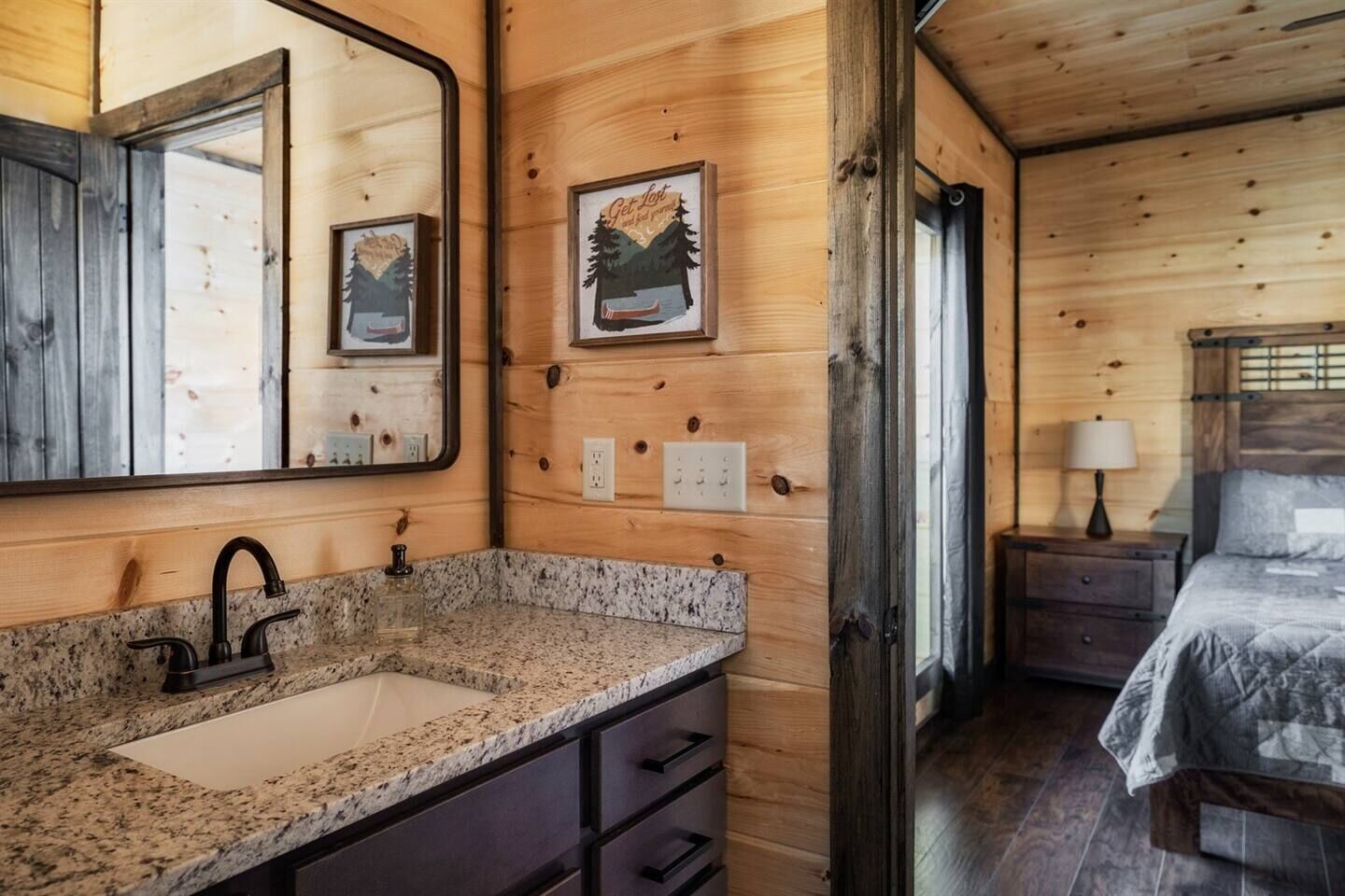 A wooden cabin interior with a granite countertop sink, a bed with a gray comforter, and two framed pictures on the wall.