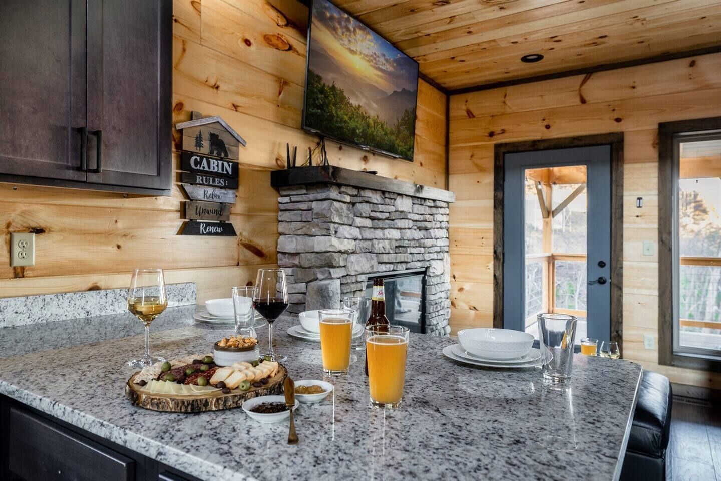 Interior de una cabaña de madera con chimenea de piedra, encimeras de granito y comedor con una tabla de quesos.