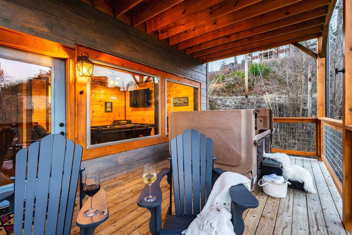 A wooden deck with two Adirondack chairs, a table with wine glasses, and a view of a billiards room through large windows.