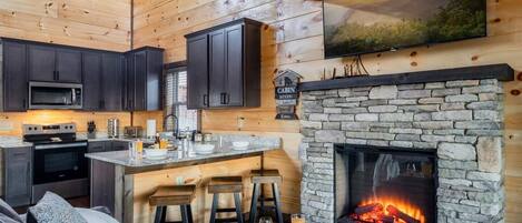 A cozy cabin interior with a stone fireplace, dark wooden cabinets, a kitchen area, and a bar with stools.
