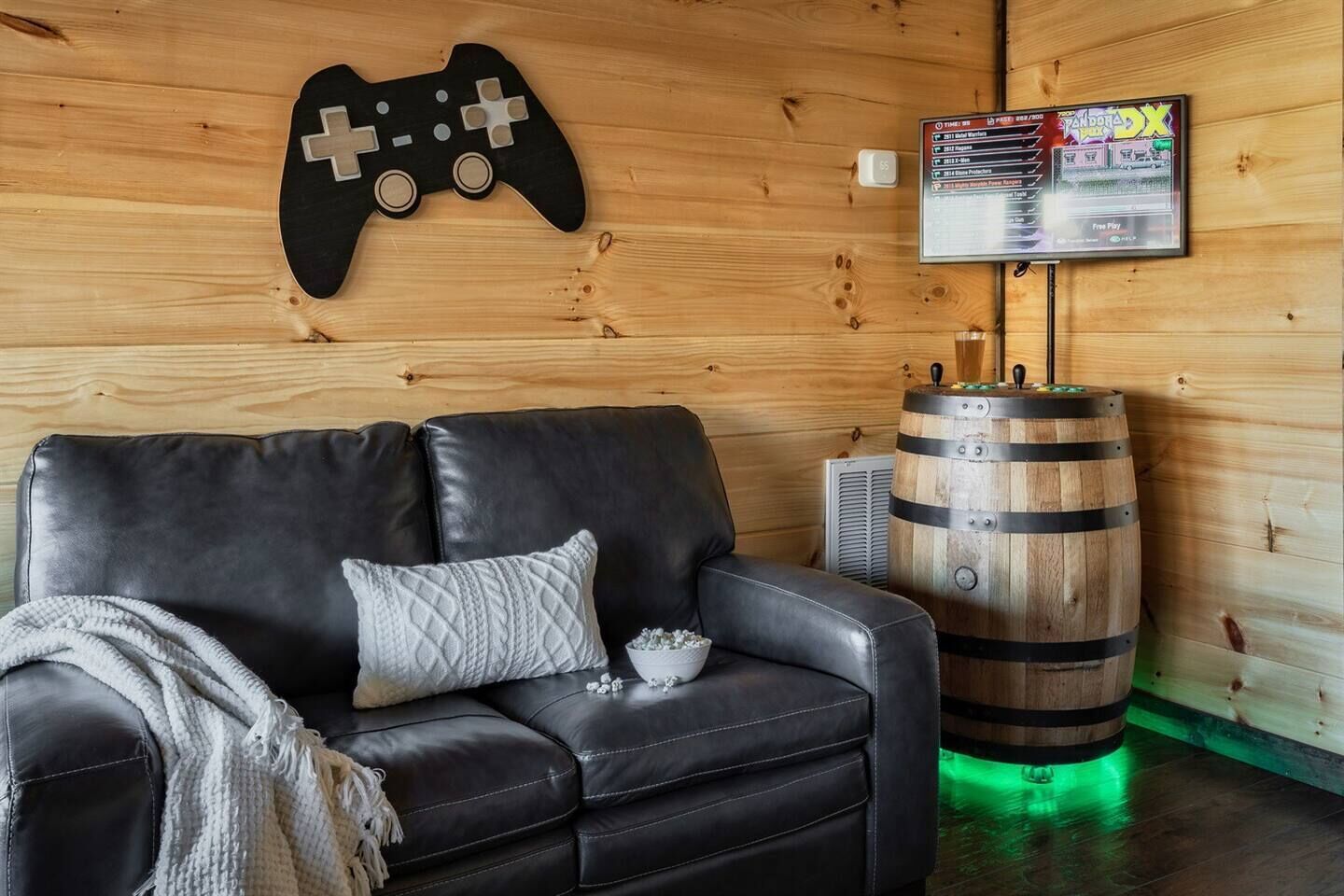 A cozy room with a black leather sofa, a wooden wall, a mounted game controller, a television, and a wooden barrel.