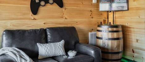 A cozy room with a black leather sofa, a wooden wall, a mounted game controller, a television, and a wooden barrel.