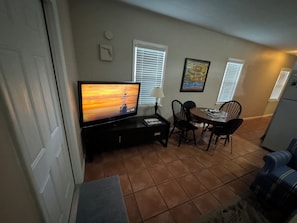 A compact living space with a TV on a stand, a dining table with chairs, a small refrigerator, and a window with blinds.