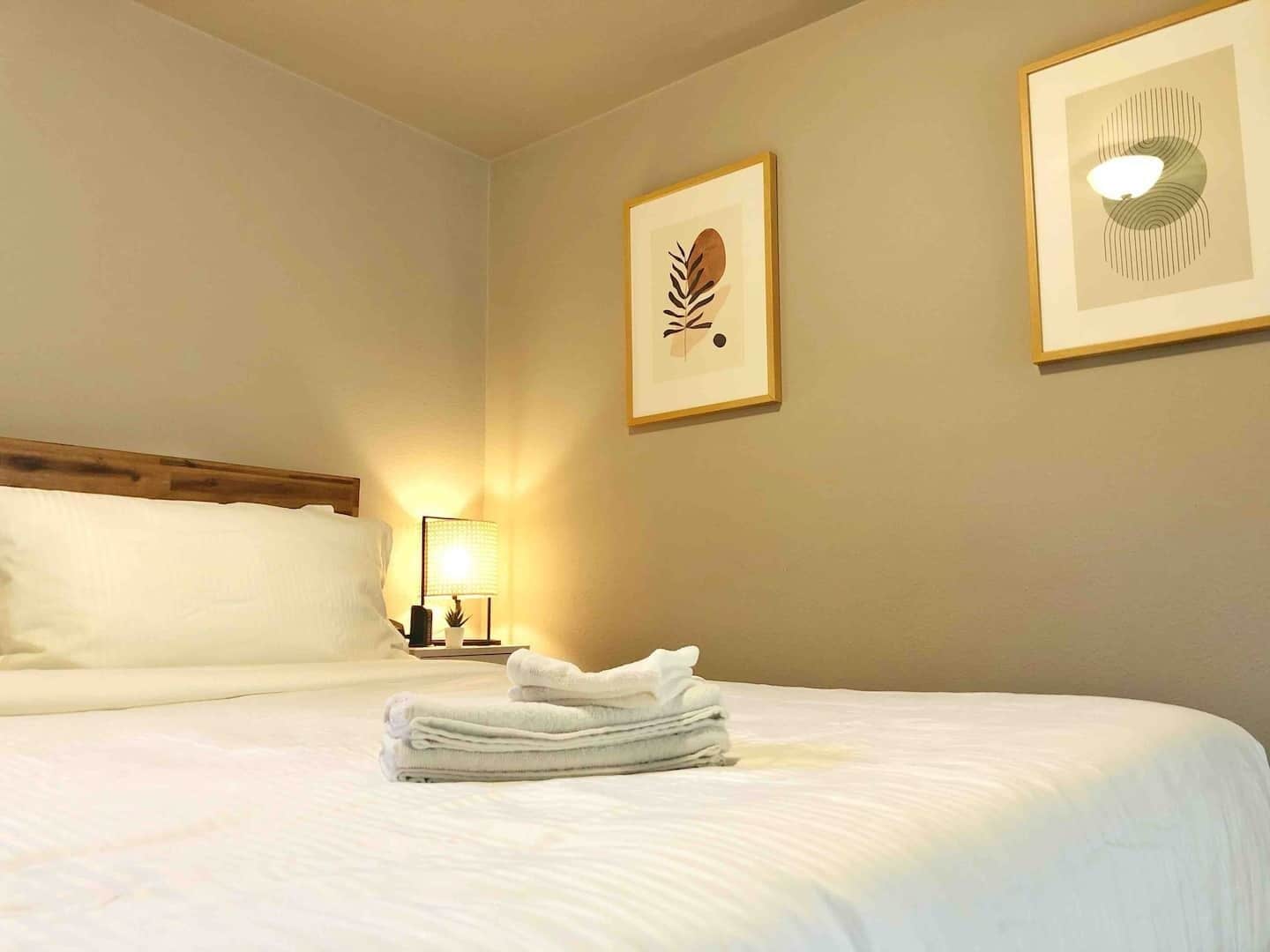 A hotel room with a neatly made bed, a bedside lamp, and two framed artworks on the wall.