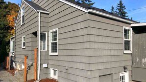A two-story house with a grey siding exterior, a front porch with a wooden railing, and a small front yard with mulch. - Seattle Launchpad - Cozy and Modern 2 BR Guest Suite (Seattle)