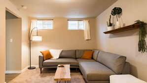 A modern living room with a grey sectional sofa, a wooden coffee table, and a white armchair. There are two windows with white curtains, a floor lamp, and a shelf with plants. - Seattle Launchpad - Cozy and Modern 2 BR Guest Suite (Seattle)