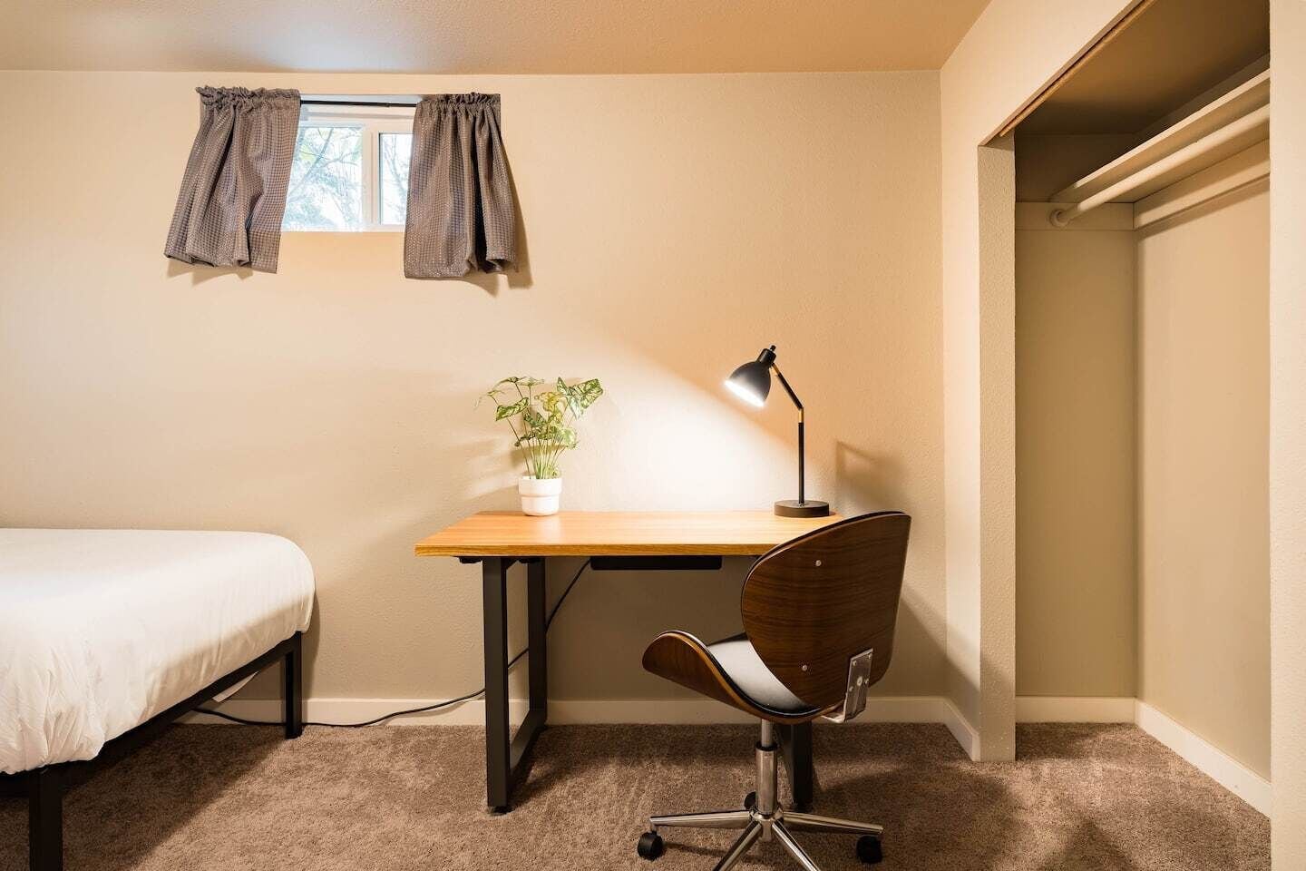 A small, neatly organized bedroom with a single bed, a desk with a chair, a potted plant, and a window with curtains.