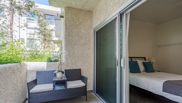 A balcony with a wicker chair and a small table, a sliding glass door leading to a bedroom with a bed and pillows.