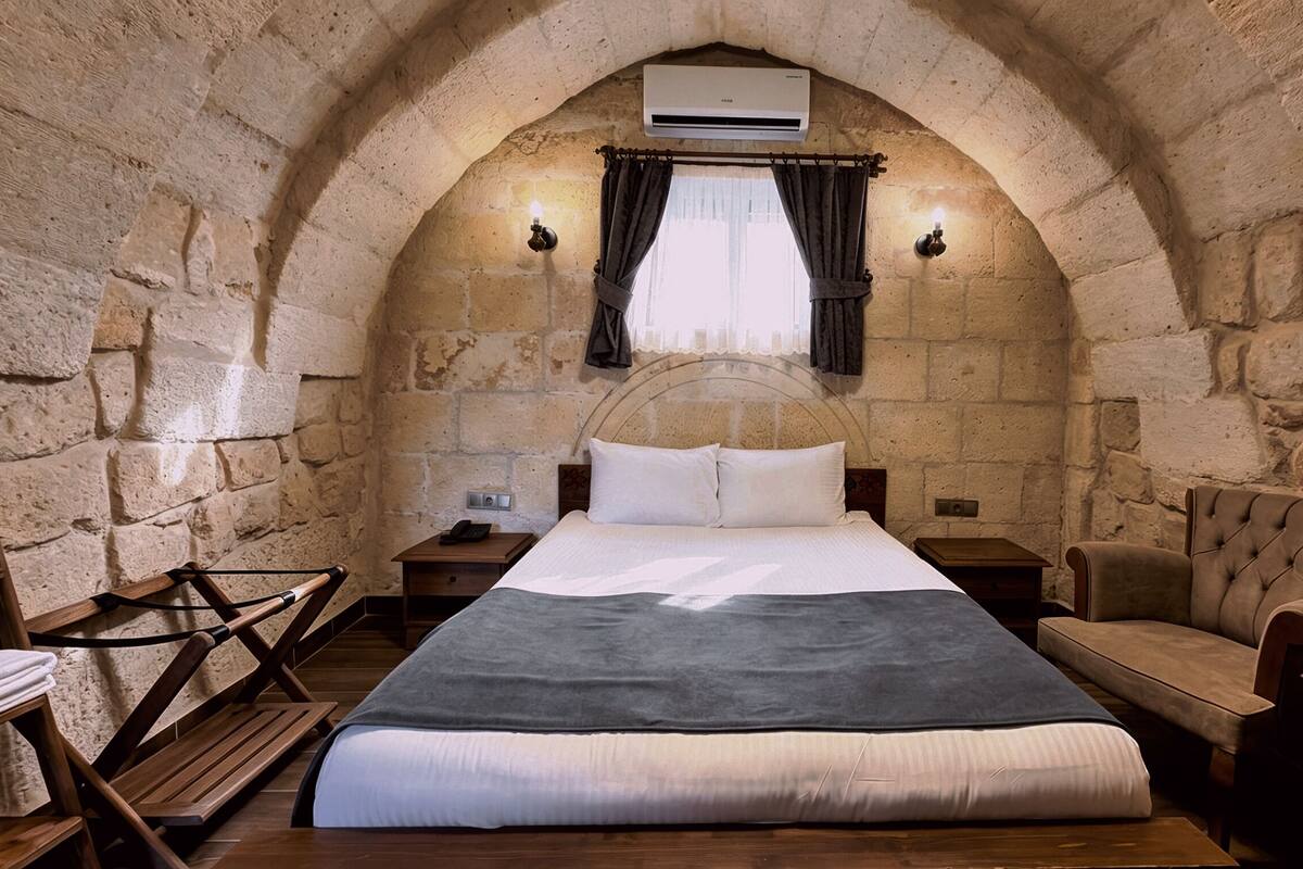 Ein Zimmer mit Steinwänden, einem Bett, einem Sessel, einem kleinen Tisch und einer Klimaanlage.