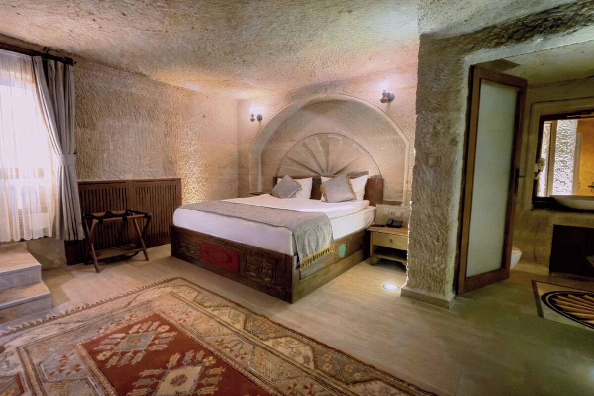 Ein rustikales Schlafzimmer mit Steinwand, einem hölzernen Bett, einem Nachttisch, einem roten Teppich mit Muster und einem Fenster mit Vorhängen.