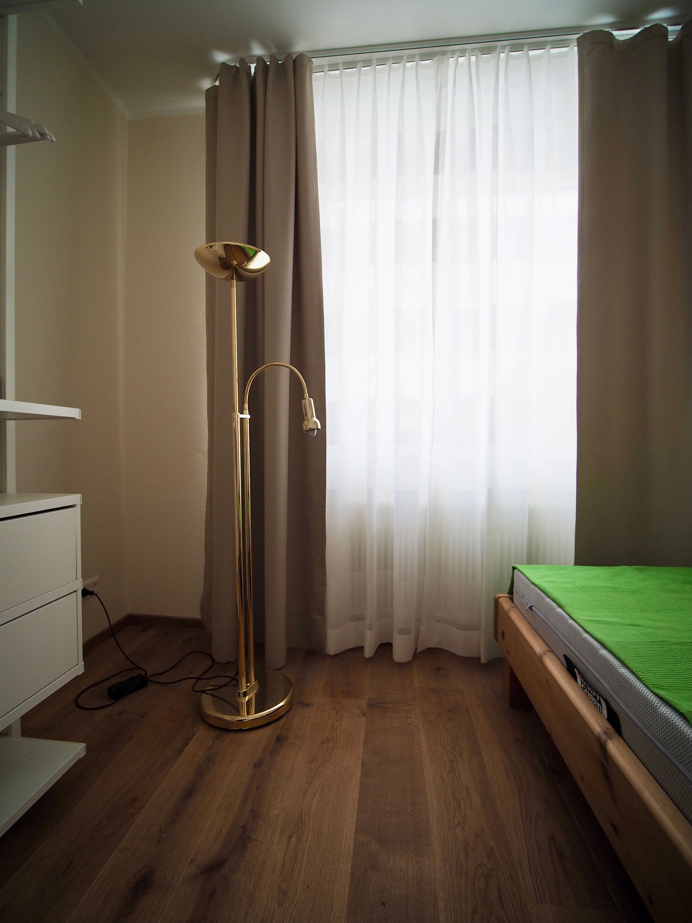 Een slaapkamer met een bed, een vloerlamp, een kaptafel en een raam met gordijnen.