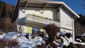 Une maison à deux étages avec un balcon, entourée d’un paysage enneigé et d’arbres.