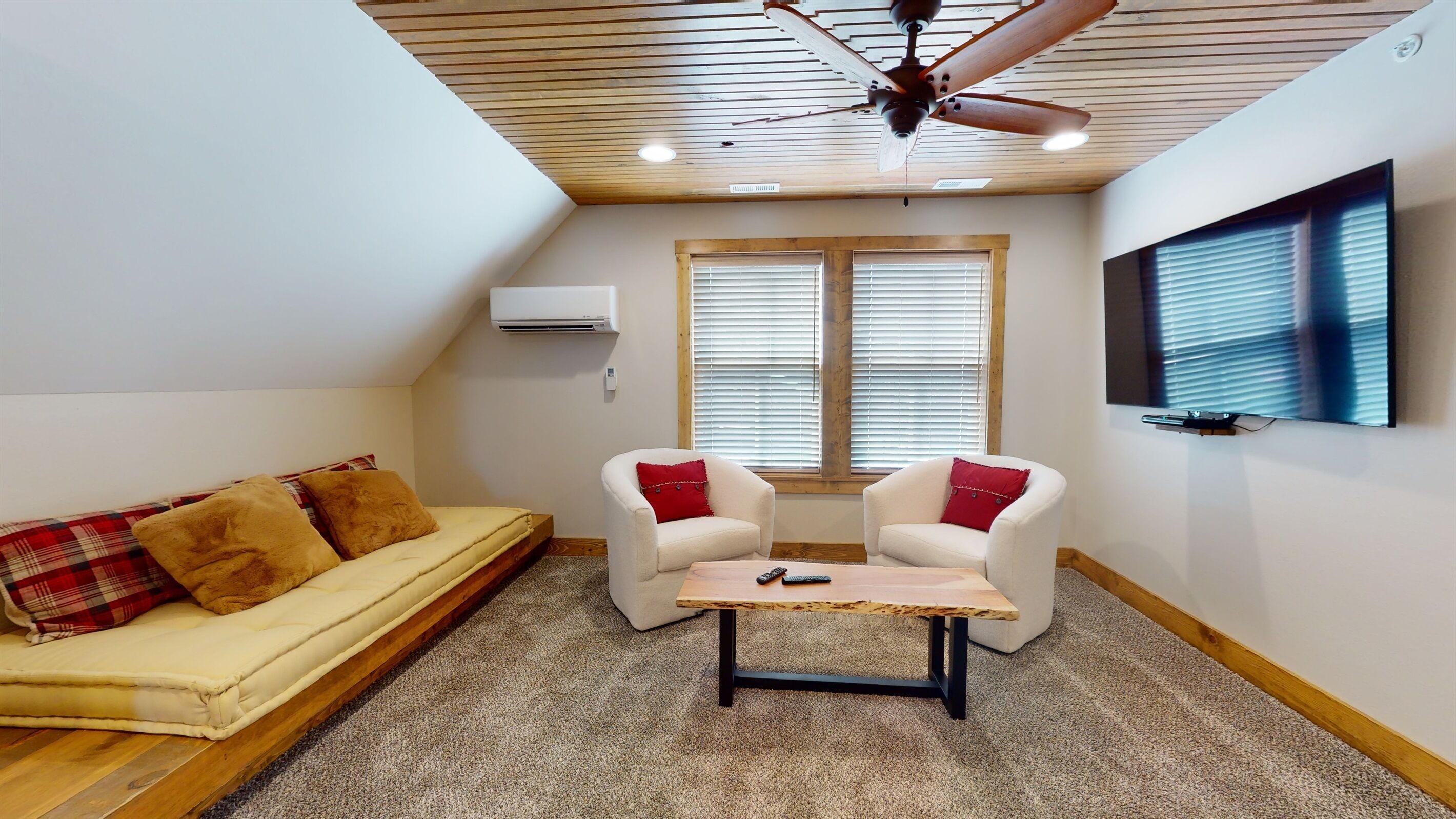 A small, cozy room with a ceiling fan, a sofa with plaid cushions, two armchairs with red cushions, a wooden coffee table, and a mounted TV on the wall.