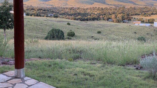 A view of a grassy field with a stone pathway leading to a wooden structure, overlooking a hilly landscape with scattered buildings.