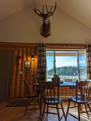 A wooden interior with a large window, a mounted deer head, and a dining table with chairs.