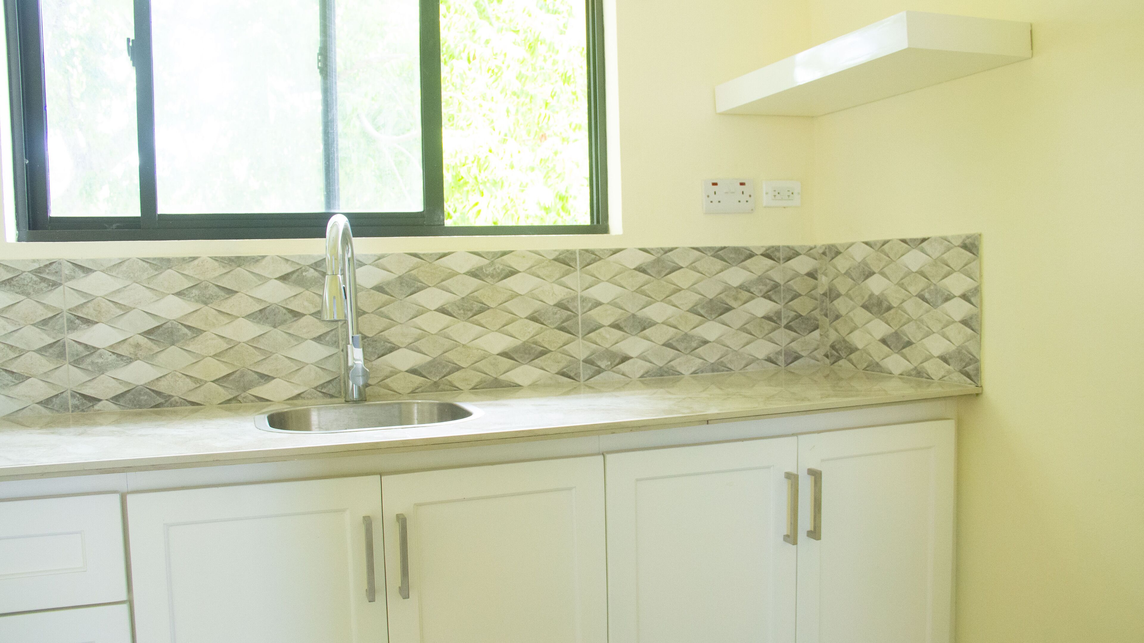 Una cucina con lavello, mobili bianchi, schienale decorato e una finestra vista verde.
