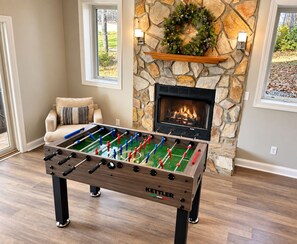 A foosball table in a room with a stone fireplace, a sofa, and a view through a window.