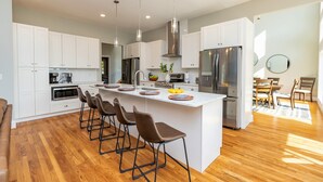 Una cocina moderna con gabinetes blancos, una isla grande con taburetes de bar, electrodomésticos de acero inoxidable e iluminación colgante.