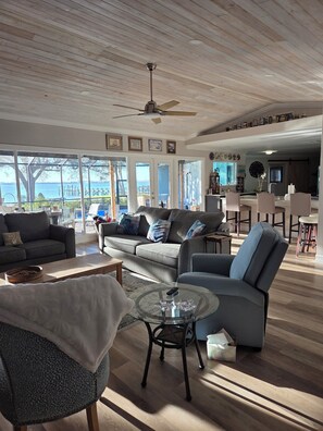A spacious living room with a large window overlooking a body of water, a ceiling fan, and a kitchen area visible through an open door. - Peace River Beach House (Port Charlotte)