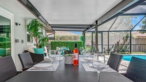 Un patio al aire libre con una mesa lista para comer, rodeado de sillas de mimbre, una botella de vino y una piscina al fondo.