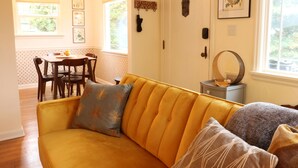 Un sofá amarillo con cojines estampados, una mesa de comedor con sillas y una ventana con vistas al verdor.