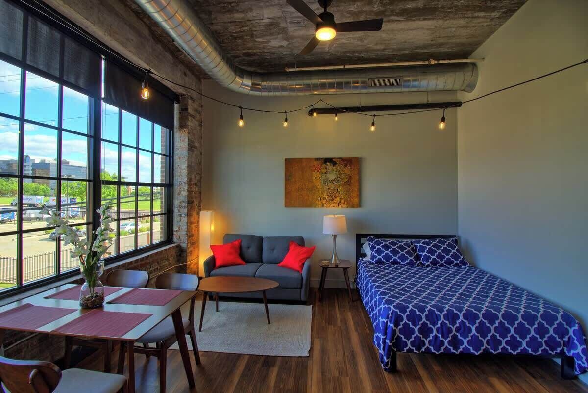 A room with a large window, a ceiling fan, a bed, a sofa, a table, and a lamp.