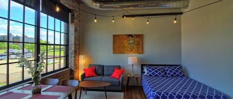 A room with a large window, a ceiling fan, a bed, a sofa, a table, and a lamp.