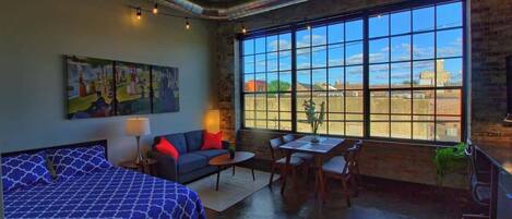 A room with a bed, a sofa, a table, and chairs, a large window with a view, and a painting on the wall.