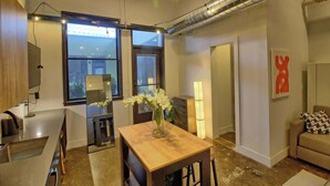A modern living space with a kitchen, dining area, and seating area. There is a large window, a ceiling fan, and a visible ductwork. - Chic Ultramodern Minimalist Studio Loft + Patio (Saint Louis)