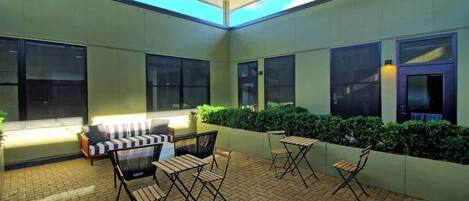 Une terrasse extérieure avec un sol en briques, plusieurs espaces de détente comprenant un canapé et des fauteuils, et de grandes fenêtres laissant entrer la lumière naturelle.