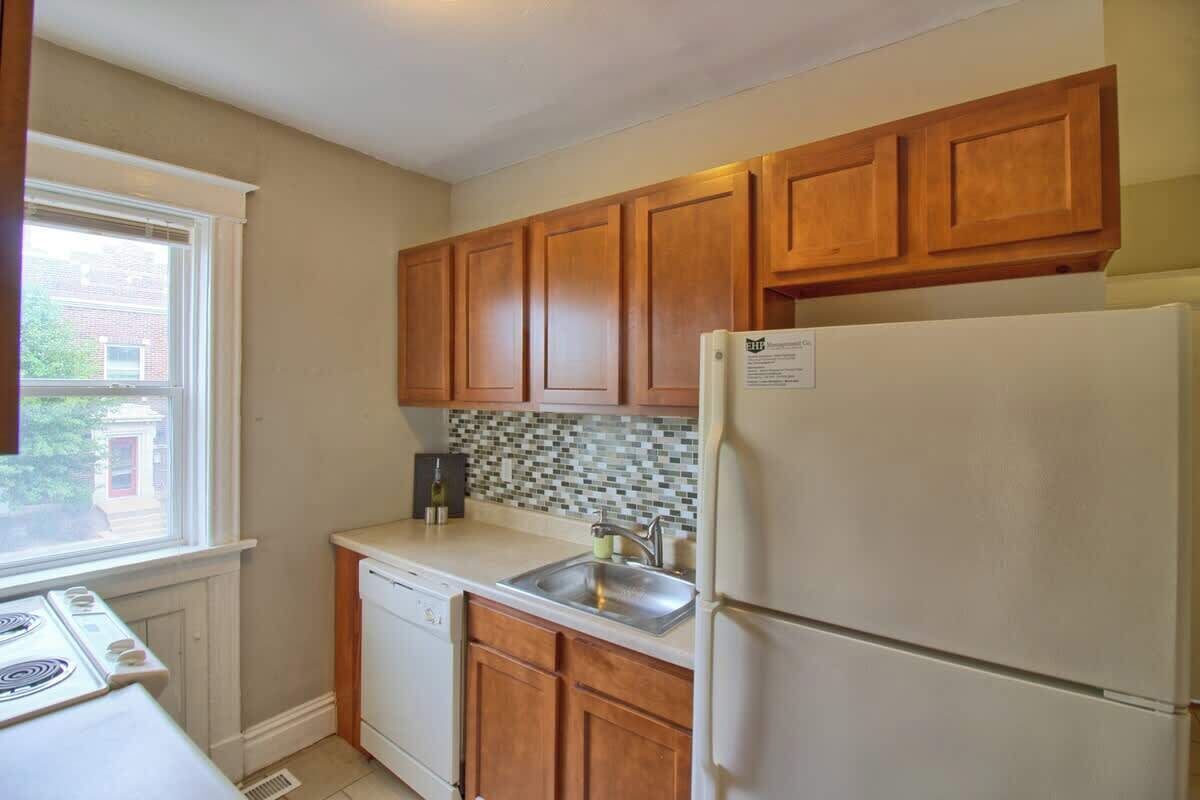 A kitchen with wooden cabinets, a white refrigerator, a dishwasher, and a window with a view of a brick building.