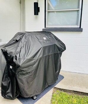 Un barbecue, recouvert d’une bâche de protection, est placé sur une terrasse en béton, à côté d’un mur blanc percé d’une fenêtre.