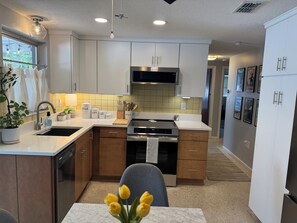 A modern kitchen with white cabinets, a stainless steel stove, and a sink. There is a dining area with a table and chairs, and a plant on the counter.