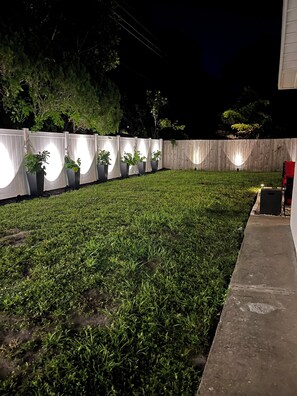 Un jardin bien éclairé, avec une clôture en bois, des plantes en pot et un chemin en béton.