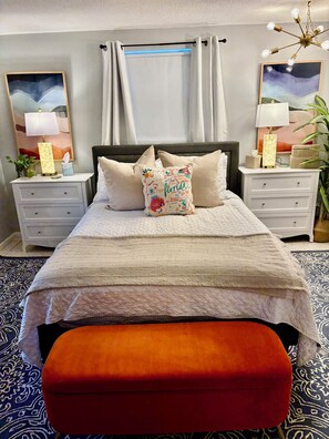 A bedroom with a bed, nightstands, a bedside table, a lamp, a plant, and a window with curtains.