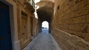 Une ruelle étroite, pavée de pierres, avec une porte bleue sur la gauche et un mur de pierre sur la droite, qui mène à un espace plus lumineux au-delà d’une arche.