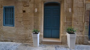 A stone building with a blue door, two white planters with plants, and a window with blue shutters. - 7 La Sengle - Valletta Dream Suites (Il-Belt Valletta)