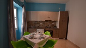 A dining area with a table set for two, two green chairs, a kitchen with wooden cabinets and a tiled backsplash, and a window with curtains. - 7 La Sengle - Valletta Dream Suites (Il-Belt Valletta)