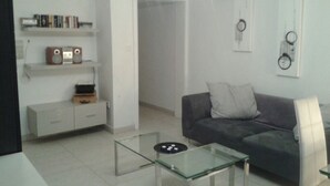 A modern living room with a grey sofa, a glass coffee table, a white cabinet, and a bookshelf with books and a radio. - Water Front Flat - balcony (Tas-Sliema)