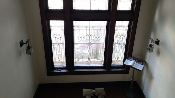 Una habitación con un gran ventanal con vidrieras y vistas al exterior. Hay una lámpara de escritorio en el lado derecho de la ventana.