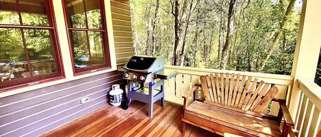 A covered porch with a grill, wooden chairs, and a view of a forest.