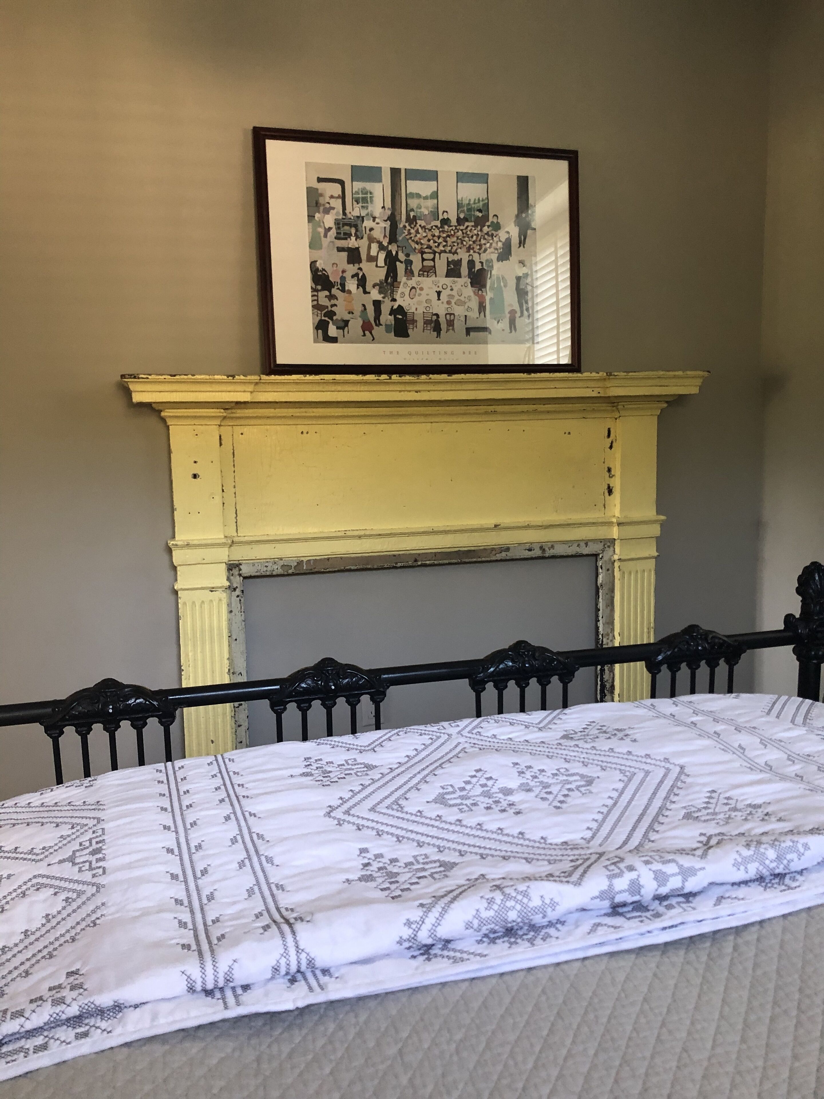 Um quarto com uma cama, uma lareira amarela e um quadro emoldurado acima da lareira.