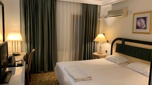 A hotel room with a bed, a TV, a desk with a lamp, a chair, and a large window with curtains. - Tas Saray Bardakci Hotel (Kahta)