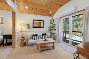 Una sala de estar acogedora con techo de madera, un sofá con cojines estampados, una mesita de centro, una lámpara y vistas a través de un ventanal corredero.