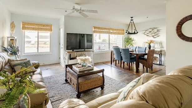 A spacious living room with a sofa, coffee table, and TV. There is a dining area with chairs and a table. - Captain’s Suite Beach Condo Near Rudee Inlet (Virginia Beach)