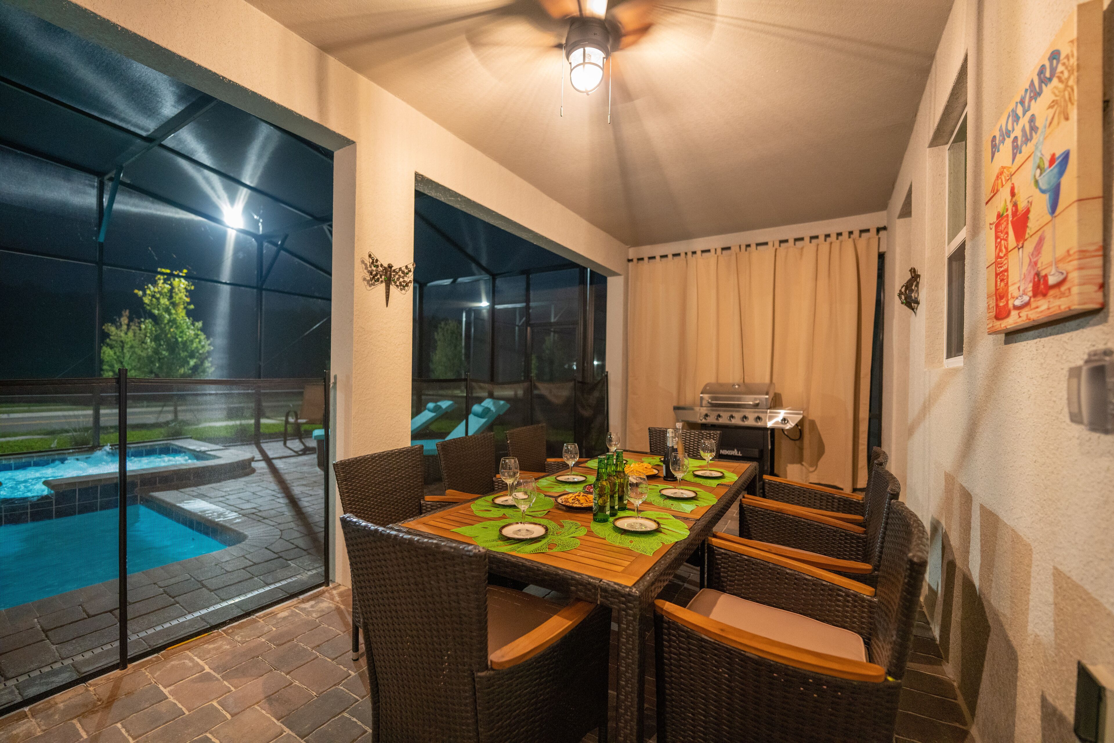 Eine Außenpatio mit Esstisch, gedeckt für sechs Personen, umgeben von Korbstühlen. Ein Pool ist durch eine Glasveranda sichtbar, und ein Grill steht in der Nähe.