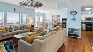 A spacious living room with a large sectional sofa, a ceiling fan, and a kitchen area with white cabinets and a microwave.