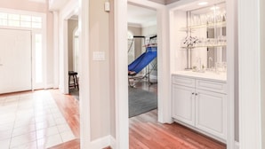 A spacious hallway with a white door, a staircase, and a kitchen area with white cabinets and a countertop.